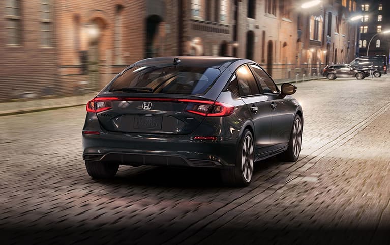 2027 Honda Civic Hatchback Sport Touring Hybrid in Meteorite Gray Metallic back driving on a cobblestone city street (Compact Hatchback).